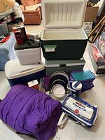 Camping gear arranged showing large and small coolers, purple sleeping bag, dishes, cooler bag, and electrical appliances.