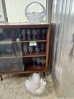 Wide view showing large crystal punch bowl on top of wooden cabinet, with multiple shelves of cut crystal stemware and additional glassware inside, and assorted decorative glass pieces on the floor in front.