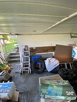 Photo showing two aluminum ladders, two tires, scrap wood panels, and tarps stored in a carport.