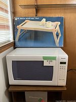 Panasonic microwave with foldable beech wood bed tray box on top, placed near window with blinds.