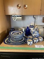 Overview of Blue Willow china set including stacked plates, cups, a teapot, and additional blue and white decorative pottery on kitchen counter.