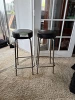 Side view of two vintage bar stools showing black round cushioned seats and chrome frames with footrests, placed on carpet near glass doors.