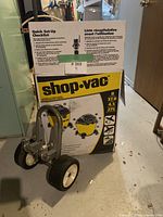 Photo of boxed Shop-Vac 8 gallon wet/dry vacuum with a metal wheel set stand placed in front of it.
