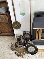 Photo showing several vintage brass kettles, a brass wall hanging, a black and gold decorative tray, pairs of brass candle holders, lantern, and smaller brass items grouped on carpet beside a fireplace.