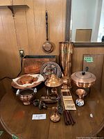 Photo showing assortment of copper and copper-plated trays, pots, chopsticks, incense burner, kettle, container, bowls, candlestick, and ceramic decorative item on wooden table.