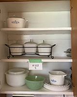 Full cabinet shelf showing vintage CorningWare and Glassbake collection with blue Cornflower pattern, mix of baking dishes and storage containers including a green glass bowl and a white measuring cup.