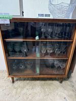 Front view of the vintage bar cabinet showing glass front sliding doors and shelves inside with glassware.