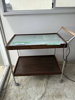 Side view of the vintage Salton electric hot cart with wooden handle and warming tray on the top shelf.