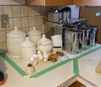 Photo showing three white ceramic canisters with lids, three stainless steel canisters, three cylindrical white ramekins, two ceramic duck figurines, three plastic jar openers, and a small honey pot arranged on kitchen counter.