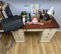 Desk with black plastic carrying case closed on left side, assorted office supplies and pink desk lamp on top