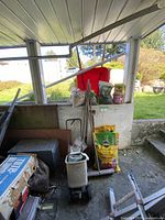 Photo showing red wheelbarrow, gardening tools, bags of soil and fertilizer stacked by a concrete wall under a carport.