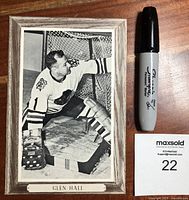 Front side of the black and white cardboard hockey photo showing Glen Hall in goalie equipment, with a Sharpie marker and auction lot card for scale.