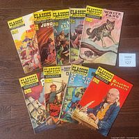 Lot of 9 Classics Illustrated comic books arranged in two rows on wooden floor, showing various colorful illustrated covers with characters, animals, and historical scenes