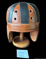 Front view of the brown and black leather helmet on a custom wooden stand against a black background.