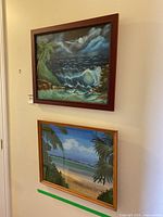Both acrylic paintings on canvas hanging on wall. Top is ocean scene with dark carved frame. Bottom is summer beach scene with lighter wood frame.