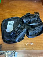 Photo showing both Bushmaster 1025 binoculars and an older pair in soft cases on wooden surface.