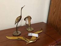 Three brass bird sculptures displayed on wooden surface: one lying flat pelican and two standing birds