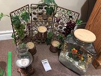 Photo showing wrought iron trellis decorated with green and purple artificial grape clusters, leaves, and three ornate candle holders with detailed bases; large glass jar with cork lid and realistic fish figurines inside simulating swimming movement when jar is moved.