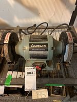 Front view of Delta bench grinder mounted with two grinding wheels and safety guards, with power cord coiled behind.