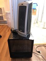 Lasko ceramic space heater standing on top of a small black electric fireplace in a room with wooden flooring and doorway in background.