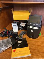 Kodak Scanza digital film scanner with its original yellow box, power adapter, cables, user manual, and film holder arranged on a wooden table.