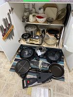 Wide view showing kitchen knives on magnetic strip, pots, pans, and kitchen gadgets inside cabinet and on floor