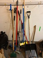 Photo showing a collection of garden and cleaning tools including shovels, rakes, broom, and squeegee leaning against a wall.