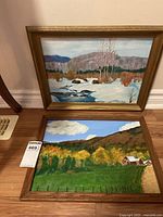 Two framed landscape paintings side by side on wooden floor near baseboard, showing winter and autumn scenes