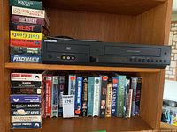 Front view of Samsung DVD/VHS combo player on shelf with assorted VHS movie tapes above and below it.