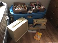 Singer Stylist zigzag sewing machine with hard case and blue bin containing sewing notions on floor next to bed.