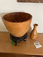 Full view of large smooth polished redwood bowl on dark ornately carved wooden stand, with small wood turned vase beside it.