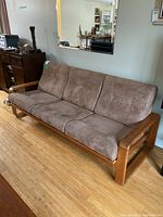 Front side view of teak framed sofa with three light brown loose cushions, showing the wooden armrest and webbed seat base.
