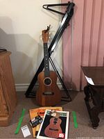 Ukulele upright next to a black metal music stand with music books arranged on the floor.