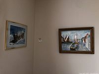Two framed watercolour paintings hanging on a beige wall, showing a ship in rough sea and a village dock scene.