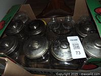 Top-down view inside cardboard box showing multiple glass jars with tarnished metal lids, varying sizes and shapes, arranged tightly.