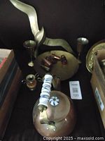 Photo of a brass tea kettle with porcelain handle and spout, small brass candlesticks, and a flat round brass decorative piece with stylized bird and twig.