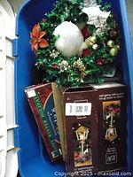 Photo shows Christmas wreath with gold, red, green ornaments, silver star, pinecones, white accents, and poinsettia flower inside blue plastic tote.