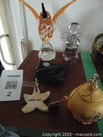 Five decorative collectible items on a wooden surface including glass hummingbird, glass angel, black glass stingray, wooden starfish keychain, and yellow hanging teapot.