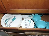 Drawer containing multiple round plates and teal-colored cups with sailing ship pattern on the plates.