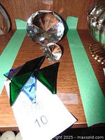 Two clear faceted glass diamond shapes—one large, one small—and a stained glass fish figurine with blue and green triangular pieces, displayed on a wood surface with green tape outlines.