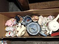 Top view of all ceramic and glass items in cardboard box showing piggy bank, salt and pepper shakers, teapots, cream and sugar set, and figurines