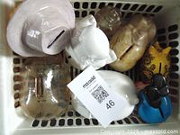 View of multiple piggy banks in a white basket including translucent, white elephant and muscular figure pigs, yellow pig character, blue bird-shaped bank.