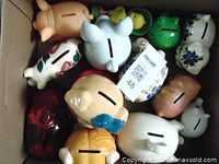 Various piggy banks in a box shown from above with coin slots visible, different shapes, colors, and paint styles.