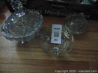 Three crystal dishes on wooden floor: large candy dish with lid and pinwheel design, small candy dish, and small bowl with feet.