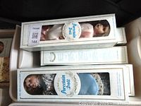 Three boxed porcelain dolls stacked inside a tote, showing different clothing colors and boxed markings