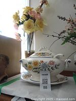 Photo of a white porcelain soup tureen with colorful floral decoration on the tureen, plate underneath, ladle inside, alongside a tall clear glass vase holding artificial flowers.