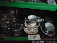 Photo showing assembled vintage tea set including glass creamer, sugar bowl with silver lid, and fine china cups stacked with saucers, displaying varied colors and floral patterns.