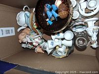 Top down view of mixed glass and ceramic items in a cardboard box showing varied shapes and colors, including white egg cups, blue ceramic birds, and glassware pieces.