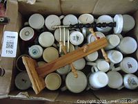 Box filled with about 30 assorted ceramic mugs, mostly white and a few with prints, plus three wooden utensils or massagers on top.
