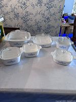 Wide photo showing set of six white CorningWare casserole dishes with matching glass lids, each featuring blue flower pattern. Arranged on table against wallpaper background.
